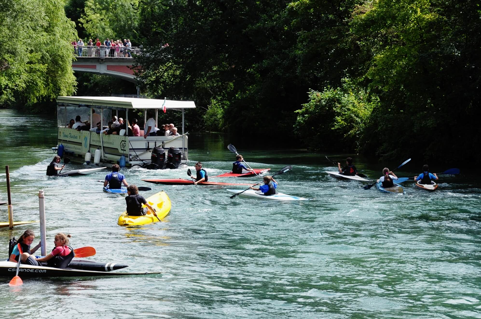 Nella foto bambini che fanno lezione di canoa sul fiume Noncello di Pordenone e persone che navigano sul battello